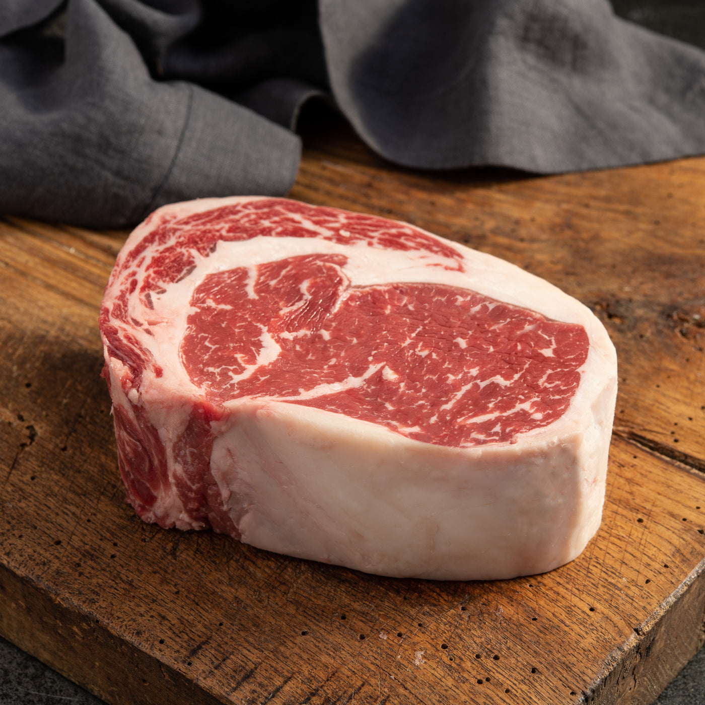 A single USDA Prime Boneless Ribeye Steak on a wooden cutting board with a dark gray cloth in the background.