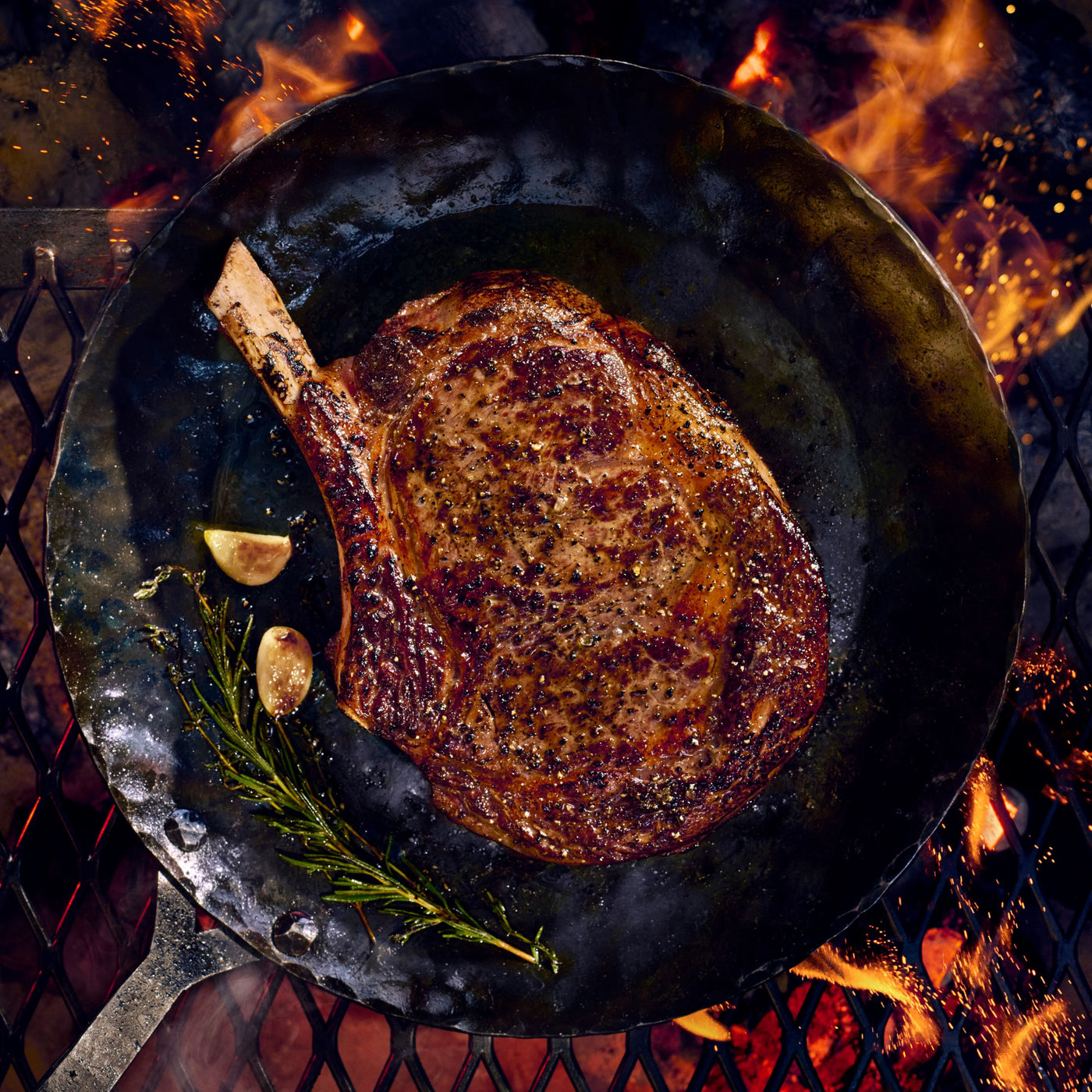 A large ribeye steak with a bone, seared to a golden-brown finish, cooking in a dark cast-iron skillet over an open fire with visible flames and embers.