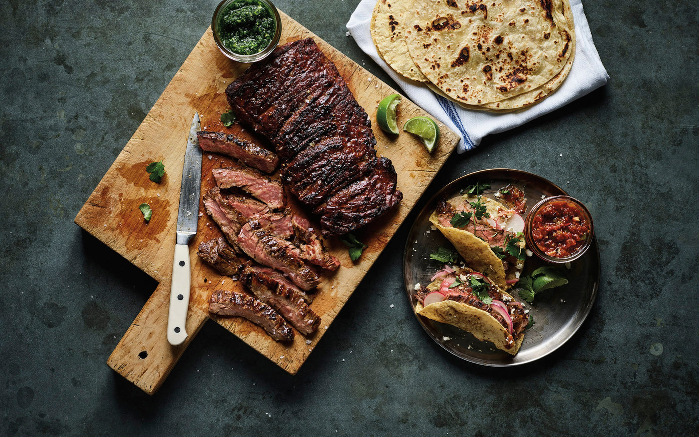 Skirt Steak on a cutting board with tortillas and sauces on a dark surface